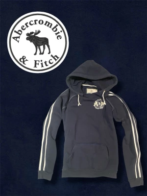 Mörkblå hoodie från Abercrombie & Fitch - Snygg mörkblå hoodie från Abercrombie & Fitch med vita ränder längs ärmarna och broderad logga på bröstet. Klassisk känguruficka framtill och justerbar huva med dragsko. Perfekt för en avslappnad och sportig stil. Står att den är storlek L men passar som S. Längd är 65cm och bredd är 47cm.