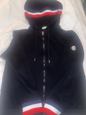 Svart Moncler hoodie med dragkedja - Svart hoodie från Moncler med dragkedja framtill och huva. Röd, vit och blå rand på huva och ärmslut. Klassisk Moncler-logga på bröstet. Mjuk bomullskänsla och snygg passform, perfekt för en stilren look.