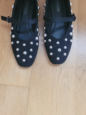 Svarta ballerinaskor med nitar från ADAMUMU - Svarta ballerinaskor från ADAMUMU med fyrkantig tå och dekorativa silvriga nitar över hela skon. Skorna har en rem över foten med spänne och är tillverkade i mockaimitation. Perfekt för dig som vill sticka ut med en edgy stil.