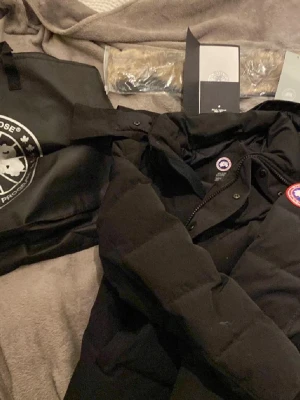  Canada Goose jacka - Säljer en svart dunjacka från Canada Goose med klassisk logotyp på bröstet. Jackan har en stilren design med hög krage och dragkedja framtill. Perfekt för kalla dagar och håller dig varm med sitt dunfoder.