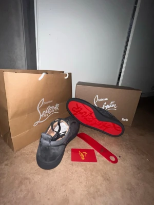 Christian Louboutin gråa sneakers - Snygga svarta sneakers från Christian Louboutin med den ikoniska röda sulan och diskret logga. Skorna har en klassisk låg modell och är tillverkade i mocka med svarta skosnören. Perfekt för dig som vill sticka ut med exklusiva detaljer.