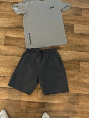 Under Armour set grå - Snyggt träningsset från Under Armour med en ljusgrå t-shirt och mörkgrå shorts. T-shirten har tryckt logga på bröstet och text på sidan, medan shortsen har elastisk midja, fickor och logga på benet. Perfekt för gymmet eller löprundan. Sitter tajt, som en size S