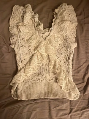 Vit spetsig stickad topp med volang - Supersöt vit stickad topp med spetsdetaljer och volang längs axlar och framstycke. Toppen har v-ringning och är ärmlös, perfekt för en romantisk look. Ribbad nederkant ger en snygg passform och det virkade mönstret gör den extra unik. 