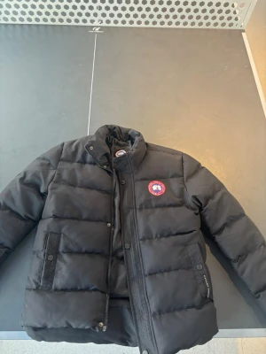 Svart dunjacka från Canada Goose - Säljer en svart dunjacka från Canada Goose med klassisk logga på bröstet. Jackan har hög krage, dragkedja och knappar framtill samt två stora fickor med lock. Perfekt för kalla vinterdagar och riktigt varm tack vare dunfyllningen.