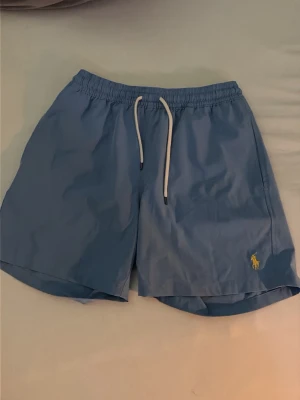 Polo Ralph Lauren badbyxor - Snygga ljusblå badbyxor från Polo Ralph Lauren med vit dragsko i midjan och klassisk gul logga broderad på vänster ben. Modellen har elastisk midja och kortare ben, perfekta för stranden eller poolen.