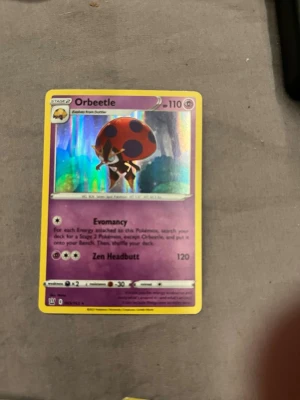 Pokémonkort Orbeetle holo - Säljer ett Orbeetle Pokémonkort med holografisk effekt. Kortet har lila och gula kanter, och visar Orbeetle med sin röda och svarta skal. Kortet har attackerna Evomancy och Zen Headbutt. Perfekt för samlare eller Pokémonfans.