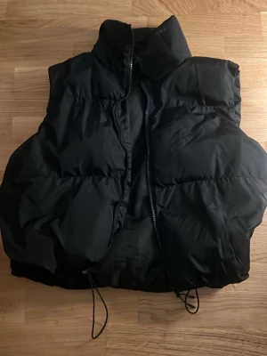 Svart puffig dunväst med hög krage - Säljer en svart dunväst med hög krage och dragkedja framtill. Västen har en puffig, oversized look och justerbar dragsko nedtill för extra stilpoäng. Perfekt att slänga över en hoodie eller tröja för en trendig lager-på-lager outfit.