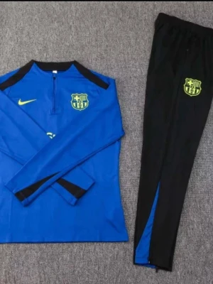 Blå Barcelona träningsset Nike - Snyggt träningsset från Nike med FC Barcelona-logga. Tröjan är blå med svarta detaljer och halv dragkedja, byxorna är svarta med blå inslag och klubbmärke. Perfekt för fotbollsträning eller chill. Materialet är troligtvis polyester för bästa komfort.