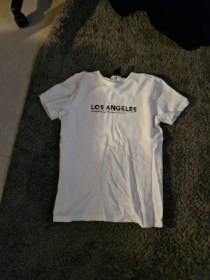 Vit t-shirt med Los Angeles-tryck - Snygg vit t-shirt med svart text 'LOS ANGELES' på bröstet. Klassisk passform och rund halsringning. Perfekt för dig som gillar enkel och stilren streetwear. Tillverkad i mjuk bomull för skön känsla hela dagen.