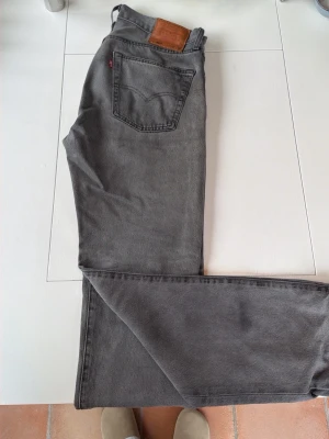 Grå Levi's raka jeans - Säljer ett par grå jeans från Levi's med klassisk femficksdesign och raka ben. Jeansen har en brun läderpatch bak i midjan och den ikoniska röda Levi's-taggen på bakfickan. Perfekta för en avslappnad och stilren look.