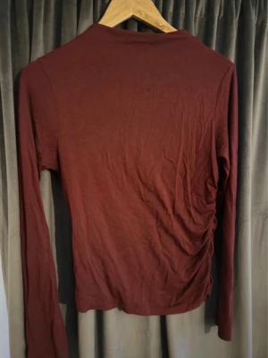 Vinröd långärmad topp med rynk - Snygg vinröd långärmad topp med rynkade detaljer på sidan som ger en cool och trendig look. Tillverkad i mjukt material som känns skönt mot huden. Perfekt att styla med jeans eller kjol för en avslappnad vibe.