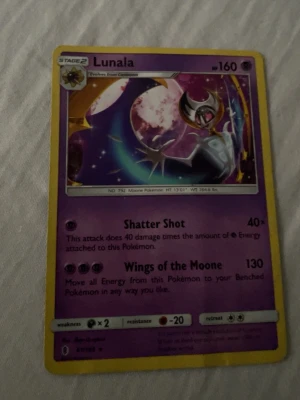Lunala Pokémonkort - Säljer ett Lunala Pokémonkort med lila och gul ram. Kortet har 160 HP och attacker som Shatter Shot och Wings of the Moone. Framsidan har en bild på Lunala mot en kosmisk bakgrund och baksidan har klassisk Pokémon-design. Perfekt för samlare eller Pokémonfans.