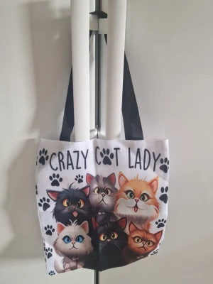 Tygväska kattmotiv kvinna - Tygväska för kvinna. Väskan är tillverkad i canvastyg och har tryck med flera katter och texten "Crazy Cat Lady" på framsidan och baksidan. Utrustad med två axelremmar i kontrastfärg. Passar för vardagsbruk, till exempel som shoppingväska eller extra vardagsväska.