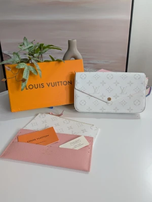 Iouis Vuitton pochette  - Detta är en elegant kuvertväska (pochette) med en slimmad och rektangulär profil. Väskan har ett lock med en distinkt V-formad skärning som stängs med en central tryckknapp. Den är tillverkad i ett ljust, belagt canvasmaterial med ett finstilt grafiskt mönster i en mjuk grå ton mot en krispigt vit bakgrund.