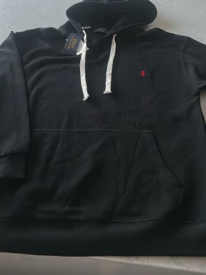 Svart hoodie från  Ralph Lauren - Svart hoodie från Polo Ralph Lauren med vit dragsko i huvan och klassisk röd logga broderad på bröstet. Hoodien har en stor magficka och ribbade muddar vid ärmslut och nederkant. Tillverkad i mjukt bomullsmaterial för en skön känsla.