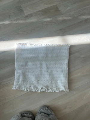 Vit smockad tubtopp med volang - Säljer en vit axelbandslös tubtopp med smockad struktur och volangkant upptill och nedtill. Perfekt till sommaren och enkel att matcha med olika stilar. Superstretchig och bekväm.