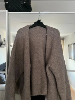 Oversize beige stickad kofta - Mysig och oversize stickad kofta i beige nyans. Koftan har långa ärmar och en öppen front utan knappar, perfekt att slänga på sig över en topp. Materialet ser ut att vara mjukt och fluffigt, vilket ger en avslappnad look.