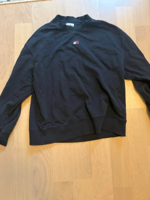 Svart sweatshirt från Tommy Hilfiger - Klassisk svart sweatshirt från Tommy Hilfiger med liten broderad logga på bröstet i rött, vitt och blått. Tröjan har rund halsringning, långa ärmar och ribbade muddar vid ärmslut och nederkant. Perfekt för en avslappnad och stilren look.