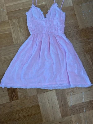 Ljusrosa kort klänning med volang - Söt ljusrosa kort klänning med smala axelband och v-ringad hals. Klänningen har volangdetaljer längs kanterna och är tillverkad i ett lätt, luftigt material som passar perfekt till varma dagar. Använd 1 gång.