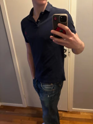 Mörkblå pikétröja från Ralph Lauren - Klassisk mörkblå pikétröja från Ralph Lauren med röd broderad logga på bröstet. Tröjan har korta ärmar, krage och knappar vid halsen. Perfekt till jeans för en clean och stilren look. 