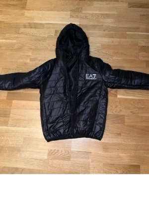 Svart pufferjacka från EA7 Emporio Armani - Snygg svart pufferjacka från EA7 Emporio Armani med huva och dragkedja framtill. Jackan har ett stilrent EA7-tryck på bröstet och quiltad design som ger en modern look. Perfekt för kalla dagar och enkel att matcha med olika outfits.