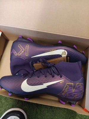 Lila Nike fotbollsskor med vita detaljer - Säljer ett par lila Nike fotbollsskor med högt skaft och vita swoosh-loggor på sidorna. Skorna har även gula grafiska detaljer och lila dobbar. Perfekta för dig som vill sticka ut på planen och ha bra grepp på gräs.