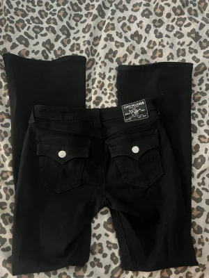 Svarta True Religion jeans bootcut - Säljer ett par svarta jeans från True Religion med bootcut-modell. Jeansen har silverfärgade knappar och detaljer, klassiska fem fickor och en patch bak i midjan. Materialet är mjukt och stretchigt för en bekväm passform. W27 använda 1 gång