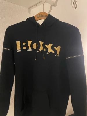Svart hoodie från BOSS med guldtryck - Svart hoodie från BOSS med stort guldigt logotryck på bröstet och dekorativa guldlinjer på ärmarna. Klassisk huva med dragsko och känguruficka framtill. Perfekt för en trendig och avslappnad stil.