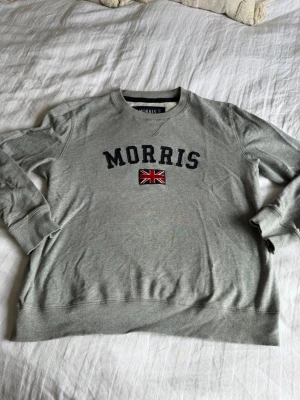 Grå sweatshirt från Morris - Snygg grå sweatshirt från Morris med broderad logga och brittisk flagga på bröstet. Tröjan har rund halsringning, ribbade muddar och är tillverkad i mjukt bomullsmaterial. Perfekt för en avslappnad och stilren look.
