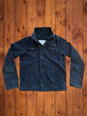 Vintage Hollister Field Jacket (sällsynt) - Säljer denna vintage, sällsynta och asfeta field jackan från Hollister. Den är riktigt sällsynt och snygg och har en unik och snygg patch över bröst fickan. Den är helt perfekt jacka nu till våren när det börjar bli varmare. Storlek är M och se måtten nedan B=66cm A=55cm