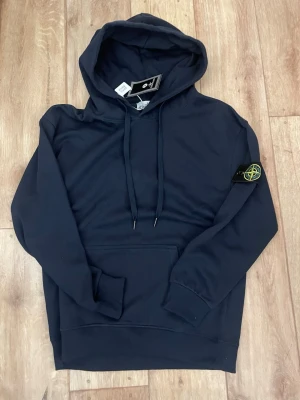 Mörkblå hoodie från Stone Island - Snygg mörkblå hoodie från Stone Island med klassisk huva och dragsko. Har en stor magficka och den ikoniska Stone Island-loggan på vänstra ärmen. Perfekt för dig som gillar streetwear och vill ha något stilrent och bekvämt.