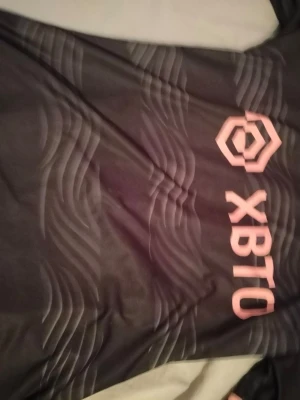 Svart sporttröja med XBTO-logga - Snygg svart sporttröja med vågigt mönster och rosa XBTO-logga på bröstet. Tillverkad i ett lätt och andningsbart material som passar perfekt till träning eller sportaktiviteter. Modern design med tryck i kontrastfärg.