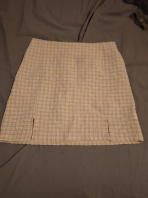 Beige rutig kortkjol med slitsar - Trendig beige kortkjol med rutigt mönster och två små slitsar framtill. Kjolen har en rak passform och är perfekt för dig som gillar en stilren och modern look. Passar till både sneakers och boots.