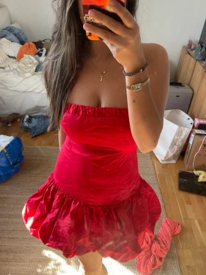 Röd axelbandslös kort klänning - Säljer en röd axelbandslös kort klänning med volangig kjol. Klänningen har en tight passform upptill och en fluffig, ballongliknande kjol. 