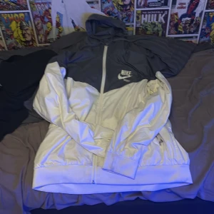 Nike vindjacka XL grå/vit - Nike vindjacka i storlek XL med huva. Jackan är tvåfärgad med mörkgrå upptill och vit nedtill, har hel dragkedja och sidofickor. Lätt och sportig känsla, perfekt för blåsiga dagar. Snygg logga på bröstet och meshfoder på insidan.