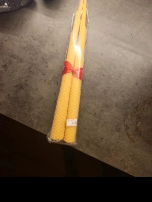 Två handrullade bivaxljus - Säljer ett paket med två smala, handrullade ljus av naturligt gult bivax. Ljusen är inslagna i plast och dekorerade med ett rött band. Perfekta för mysiga kvällar eller som present.