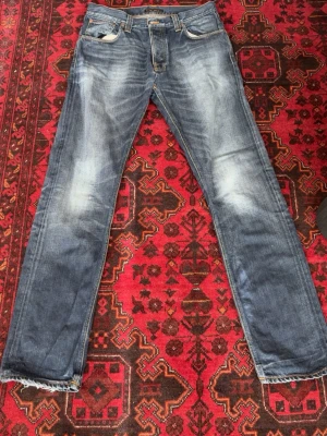 Blå slitna jeans från Nudie Jeans Hank Rey model - Säljer ett par ovanliga Hank Rey Jeans från Nudie Jeans, Jeansen går inte att köpa längre. Jeansen har slitningar och tvättade detaljer. Jeansen har raka ben, fem fickor och synliga sömmar på bakfickorna. Perfekta för en avslappnad och trendig look.