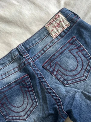 True Religion blå bootcut jeans - Säljer ett par blå jeans från True Religion med ikoniska röda sömmar och stora bakfickor. Modellen har bootcut. Jeansen är i denim med snygg tvätt och detaljerade kontrastsömmar❤️