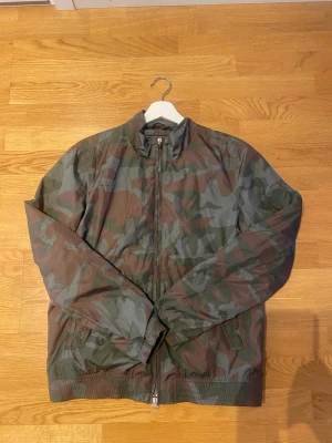 Grön camo vindjacka från Woolrich - Säljer en snygg vindjacka från Woolrich med klassiskt grönt camouflagemönster. Jackan har dragkedja fram, ståkrage och två sidofickor. Tillverkad i lätt och vindtätt material, perfekt för blåsiga dagar. Stilren och skön!