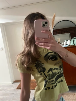 Gul croppad t-shirt med Los Angeles-tryck - Cool gul t-shirt med svart Los Angeles-tryck, palmer och bilmotiv. Croppad modell med korta ärmar och rund hals. Perfekt för dig som gillar streetstyle och grafiska prints.