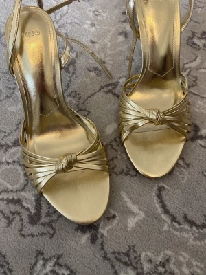 Guldiga pumps med knut från Mango - Säljer ett par snygga guldiga pumps från Mango med smala remmar och dekorativ knut framtill. Skorna har öppen tå, smal klack och justerbar ankelrem. Perfekta för dig som vill sticka ut med metallic och elegant design. Använd en gång till balen!