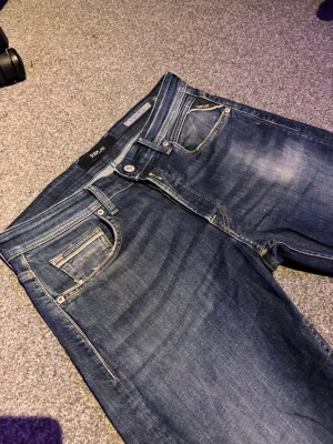 Replay Jeans - Snygga blå slim fit jeans från Replay med klassisk femficksdesign och diskreta slitningar framtill. Jeansen har en något tvättad look och silverfärgade detaljer vid knappar och nitar. Perfekta för en trendig och avslappnad stil.