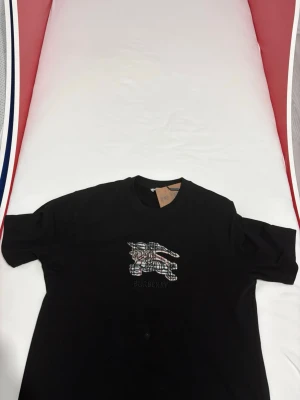 Svart Burberry t-shirt med rutig logga - Snygg svart t-shirt från Burberry med klassiskt rutigt tryck av riddare på bröstet och diskret Burberry-text. Använd endast 1 gång. PRISET KAN DISKUTERAS!