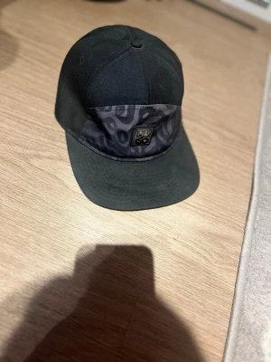 Svart keps med mönstrad panel - Säljer en svart keps med böjd skärm och en unik mönstrad panel framtill. På panelen sitter en liten patch med ett coolt motiv. Kepsen har klassisk form och passar perfekt till streetwear-stilen.