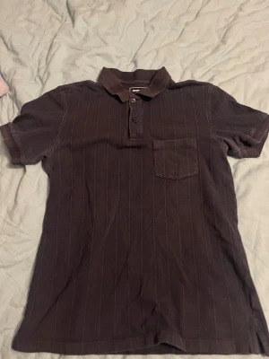 Mörkblå randig piké med ficka - Snygg mörkblå pikétröja från Threadbare med tunna vertikala ränder och klassisk krage. Tröjan har korta ärmar, knappslå med två knappar och en bröstficka. Perfekt för en avslappnad men stilren look.