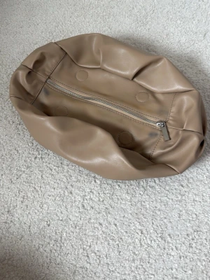 Beige mjuk handväska i fejkläder clutch - Säljer en beige handväska i mjukt fejkläder med rundad form och dragkedja på undersidan. Väskan har minimalistisk design utan synligt märke och passar perfekt till en clean och stilren look.