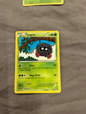 Tangela Pokémonkort - Tangela Pokémonkort med grön och gul ram, motiv av Tangela som hänger från en gren. Kortet har färgglad design med blå himmel och gröna detaljer. Materialet är laminerad papp och kortet har text på engelska samt symboler för attack och energi.
