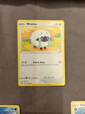Pokémonkort Wooloo - Säljer ett Pokémonkort med Wooloo-motivet. Kortet har en vit och gul ram, med en tecknad bild av Wooloo, ett får-liknande Pokémon med svart ansikte och vita lockar. Kortet är tillverkat i papper och har engelska texter och symboler.