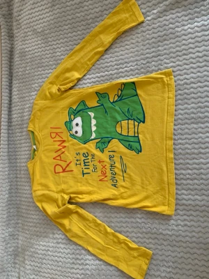 Gul långärmad topp med dinosaurie - Gul långärmad topp med en stor grön dinosaurie och texten 'RAWR It's time for the next adventure' på framsidan. Tillverkad i mjuk bomull, perfekt för en lekfull och färgglad stil.