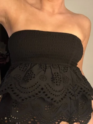 Svart bandeau tubtopp med spets - Säljer en svart bandeau tubtopp med smockad överdel och vacker spetsdetalj i flera lager. Toppen är ärmlös och har ett broderat mönster med hål och volang. Perfekt för dig som vill ha en trendig och unik topp.